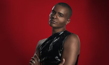 Layton Williams, a young black man with short black hair, in a leather top with two hoop earrings and two large rings stood with his arms crossed and a neutral expression looking at the camera on a red background