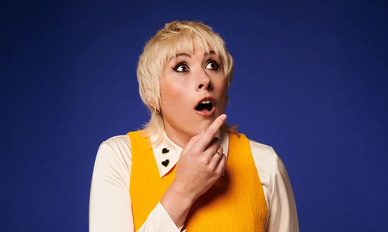 Maisie Adam, a white woman with blonde hair, in a mullet style, dressed in a white shirt with two black hearts on the collar and a yellow sweater vest. She is looking to the right shocked and pointing