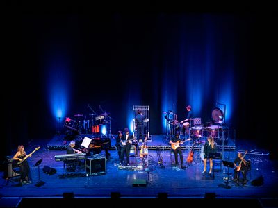 A band performs on stage under blue lighting, with musicians playing a variety of instruments. At the back of the stage, one performer plays a set of tubular bells.