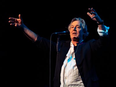 A man in a white shirt and dark jacket stands at a microphone with his arms raised and outstretched against a black background.