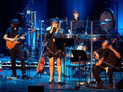 A woman sings on stage while a drummer performs behind her, with a guitarist and an electric cellist on either side. Blue stage lighting illuminates the performers, who are all dressed in black.