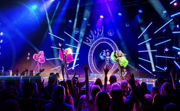 Four drag queens on stage on matching sparkly bodysuits in pink, purple, blue, and green. They're performing in front of a large Q with a crown on top in front of a very enthusiastic audience