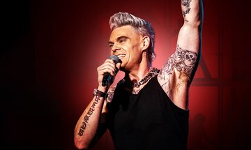 A Robbie Williams impersonator in a black vest top with lots of tattoos smiling as he holds a mic up to his mouth in one hand and has his other arm extended up above his head