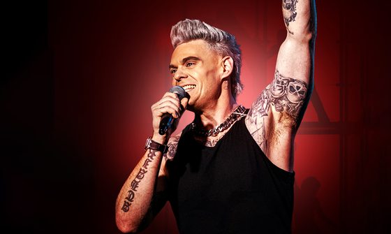 A Robbie Williams impersonator in a black vest top with lots of tattoos smiling as he holds a mic up to his mouth in one hand and has his other arm extended up above his head
