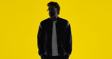 Romesh Ranganathan in low lighting in a black bomber jacket with a white shirt and black jeans on a bright yellow background