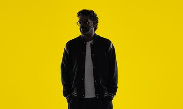 Romesh Ranganathan in low lighting in a black bomber jacket with a white shirt and black jeans on a bright yellow background