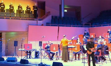 A photo taken in The Anvil of The Royal Marines Band performing on stage under purple lighting with five more members performing from the choir stalls under yellow light
