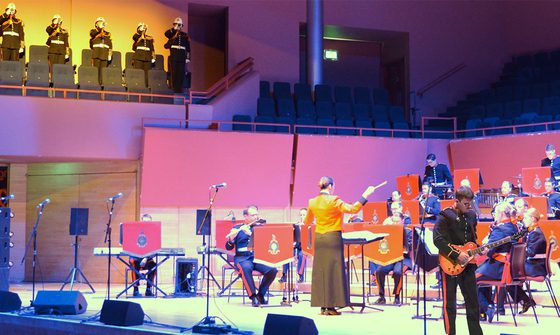 A photo taken in The Anvil of The Royal Marines Band performing on stage under purple lighting with five more members performing from the choir stalls under yellow light