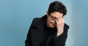 Sue Perkins, a white woman with short black hair and glasses resting her forehead on her hand with a soft smile on her face. She is wearing a black duffle coat on a light blue background