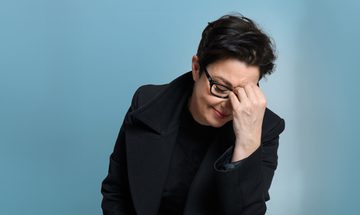 Sue Perkins, a white woman with short black hair and glasses resting her forehead on her hand with a soft smile on her face. She is wearing a black duffle coat on a light blue background