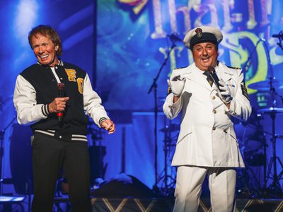 Two men smile on stage holding microphones. One wears a naval captain's uniform and the other a varsity jacket.