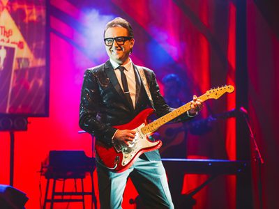 A man wearing glasses and a black sequin jacket smiles while playing the guitar.