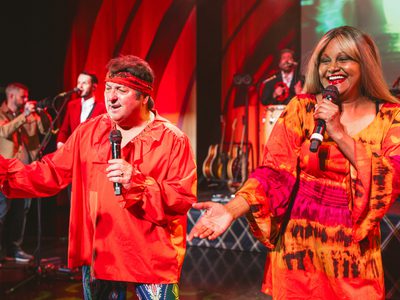 A male and female performer in orange 1960s-inspired outfits sing on stage, with a tambourine player, backing singer and photographer in the background.