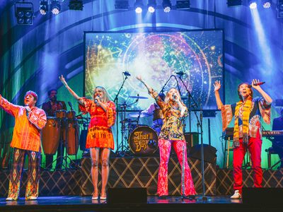 Two men and two women wearing colourful sixties inspired outfits. They are holding microphones, There is a band accompanying them in background and a colourful screen with an image of star signs.