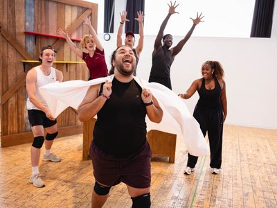 Six actors rehearsing. There is one at the front holding a sheet over his shoulders like a cape with his tongue out. Three are behind on a crate with their hands in the air, and two more either side watching the caped one and smiling