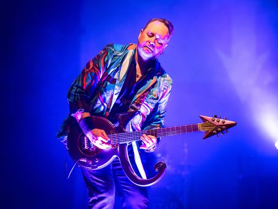 A guitarist wearing a colorful reflective jacket plays an electric guitar on stage under blue lighting.