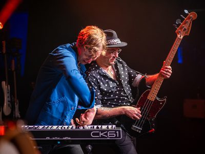 Two men stand side by side performing music. The man on the left, wearing a blue shirt, plays a keyboard, while the man on the right, wearing a black-and-white polka-dot shirt and a black hat, plays a guitar.
