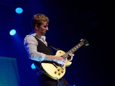 A man wearing a white shirt and black waistcoat plays a guitar on stage, illuminated by blue lights behind him.