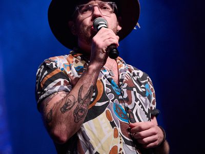 A man wearing glasses, a hat, and a colorful shirt sings into a microphone on a stage with blue lighting and mist in the background.