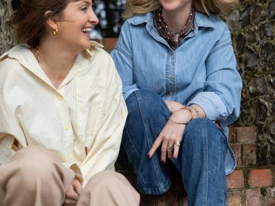 Two women sitting on a brick fence, smiling at each other as their hair blows in the wind.