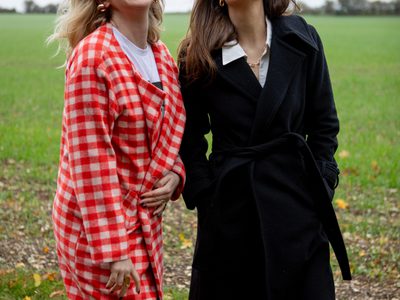 Two women standing in a grassy open space on a windy day, smiling. The woman on the left is wearing a white and red check jacket, while the woman on the right is wearing a black jacket.