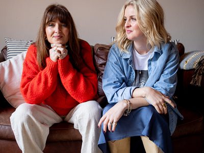 Two women sitting on a brown leather sofa; one rests her head on her clasped hands while the other sits with her arms crossed, looking at her.