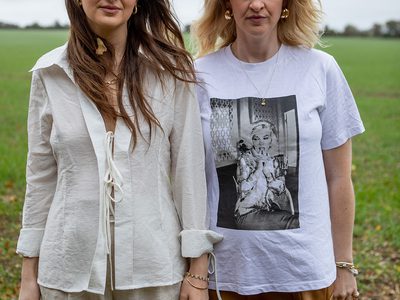 Two women standing side by side in a grassy open space, their hair blowing in the wind.