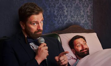 A bearded man in a dark tuxedo and bow tie sits on the edge of a bed, holding a microphone and pointing toward another bearded man lying under a pale pink blanket.