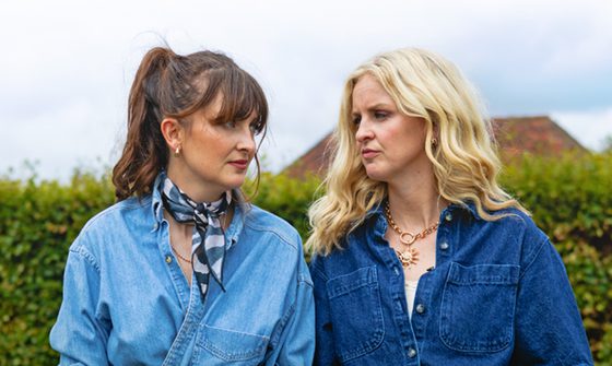A brown-haired woman and a blonde woman in denim stare at each other in front of a bush beneath a foggy sky.
