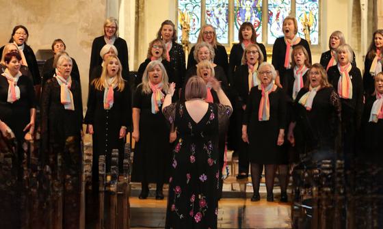 The Hampshire Harmony Ladies Choir all dressed in black with matching white and pink scarves in a church singing in front of a stained glass window