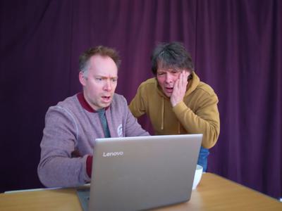Two men in casual clothes one in a yellow hoodie one in a red jacket rehearsing for the show. The man in the red jacket is sat at a laptop shocked, the other in the yellow hoodie is looking over his shoulder with his hand on his face also looking shocked.
