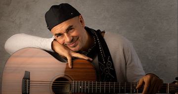 Antonio Forcione leaning on a guitar and smiling at the camera with his face resting on his hand. He is wearing a white long sleeve top and black scarf and hat
