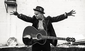 A black and white photo of Dean Owens, an older white man wearing a hat, shirt and jacket with a guitar strapped around him. He is stood in front of a white building with an American stop sign on the floor and his arms outstretched either side of him