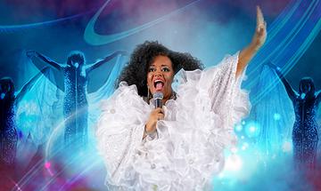 A Diana Ross Impersonator in a large ruffled white dress singing into a microphone with one arm up and other dancers in the background all in a blue light