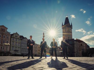 G4 dressed in dark suits, standing in Prague Square with the sun and blue sky behind them