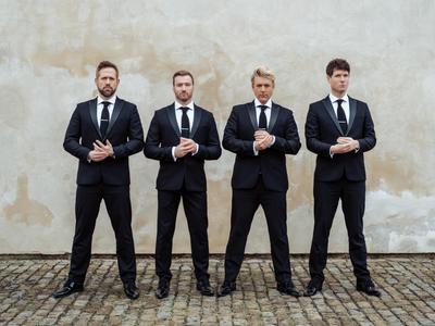 G4 dressed in dark suits, standing in front of an off-white wall with their hands clasped together and neutral facial expressions