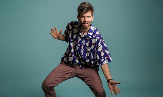 Ivo Graham wearing a blue shirt with ducks on, crouching and looking at the camera with his mouth open. He has one hand up and one hand down