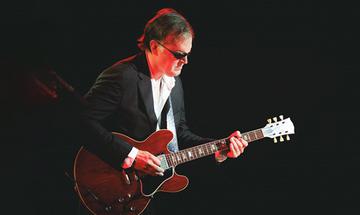 Joe Bonamassa wearing dark glasses, a white shirt, and a black blazer, playing a red electric guitar on a dark background