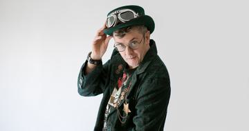 Joe Pasquale standing sideways looking over his shoulder with his hand on his hat