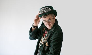 Joe Pasquale standing sideways looking over his shoulder with his hand on his hat.