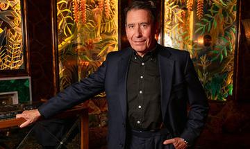 Jools Holland standing with one hand in his pocket and the other leaning on a table. He is wearing a dark suit and smiling at the camera