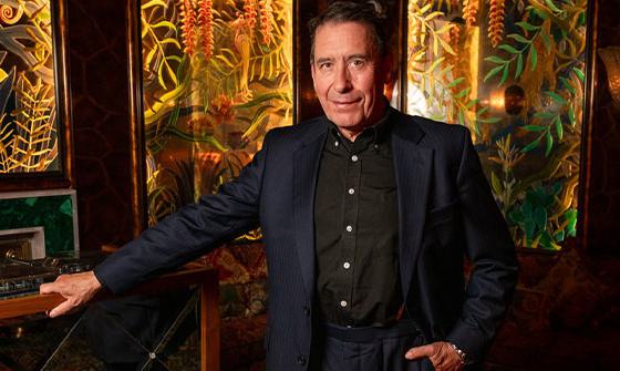 Jools Holland standing with one hand in his pocket and the other leaning on a table. He is wearing a dark suit and smiling at the camera