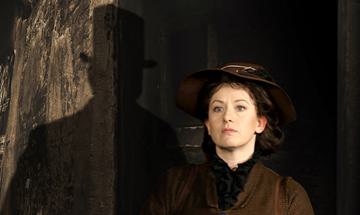 A lady wearing a brown coat stood by a doorway with a shadow of a man in a top hat next to her
