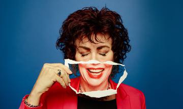 Ruby Wax wearing a red suit jacket, holding a piece of paper with a mouth on that fits with where her mouth would be