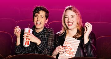 Two women sat in auditorium seats holding popcorn and a drink. They are smiling at something to the side of the camera, and there is a pink background