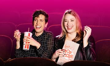 Two women sat in auditorium seats holding popcorn and a drink. They are smiling at something to the side of the camera, and there is a pink background