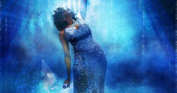 A Whitney Houston tribute on stage with her arm up in the air, looking towards it. She is wearing a blue sparkly dress on a blue-lit background
