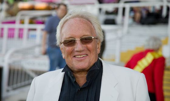 John Sutton headshot. He wears a black shirt and white jacket and is smiling at the camera