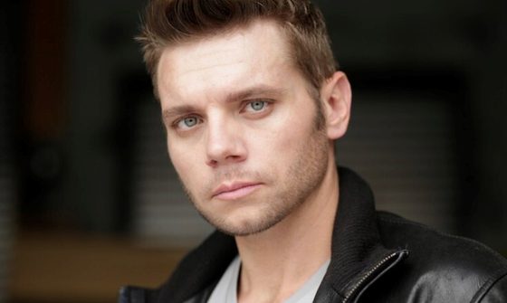 Nathaniel Hackmann, a white man, with short, quiffed, light brown hair and stubble, wearing a white t-shirt and leather jacket looking at the camera with a neutral expression