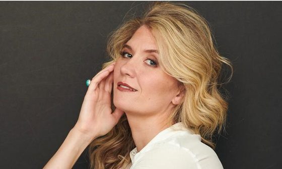 Scarlett Strallen, a white woman with blonde wavy hair, wearing blue skinny jeans and a white blouse. She is sat holding on knee with one arm and gently touching her face with the other. She is smiling softly at the camera.